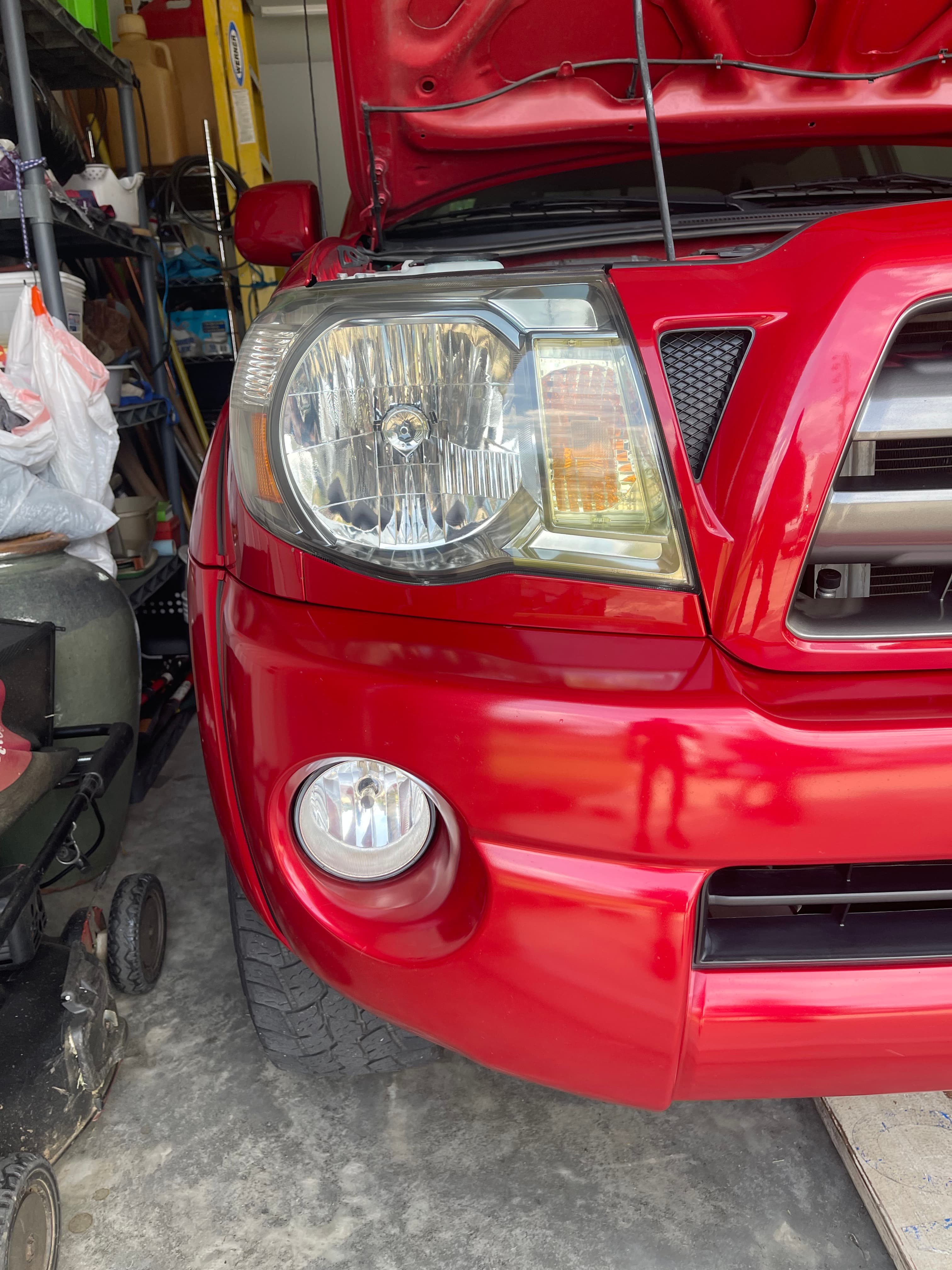 Close-up of a red vehicle's headlight and fog light assembly in a cluttered garage.
