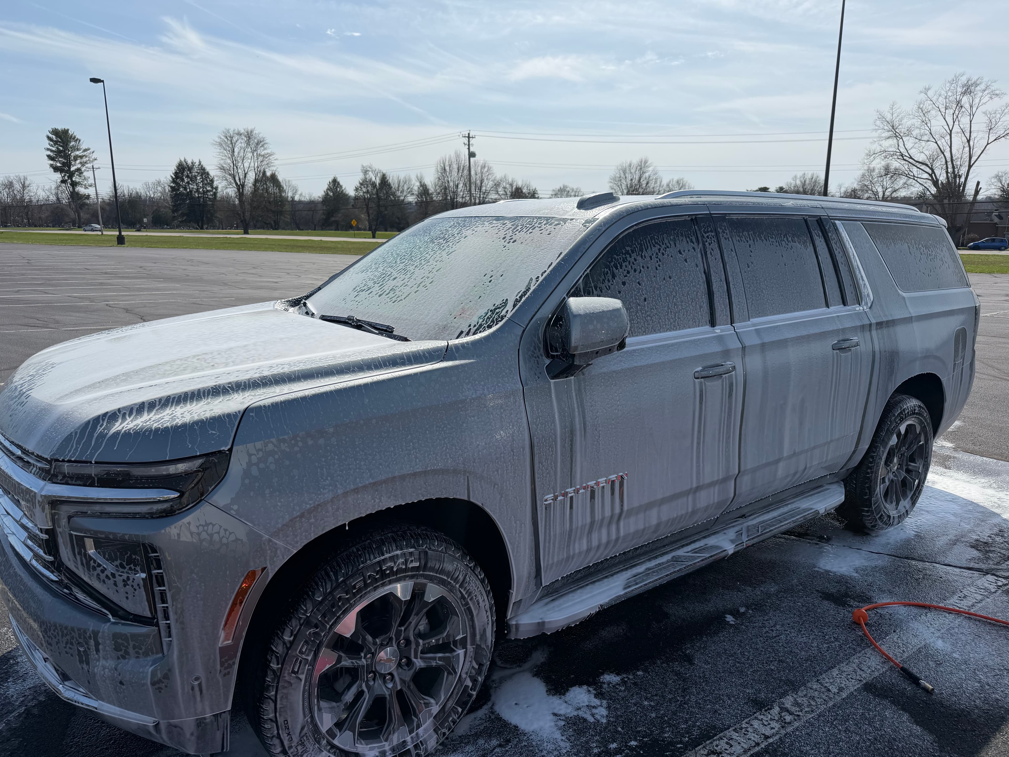 Grey Chevrolet Suburban covered in white soap suds being washed in an outdoor parking lot.